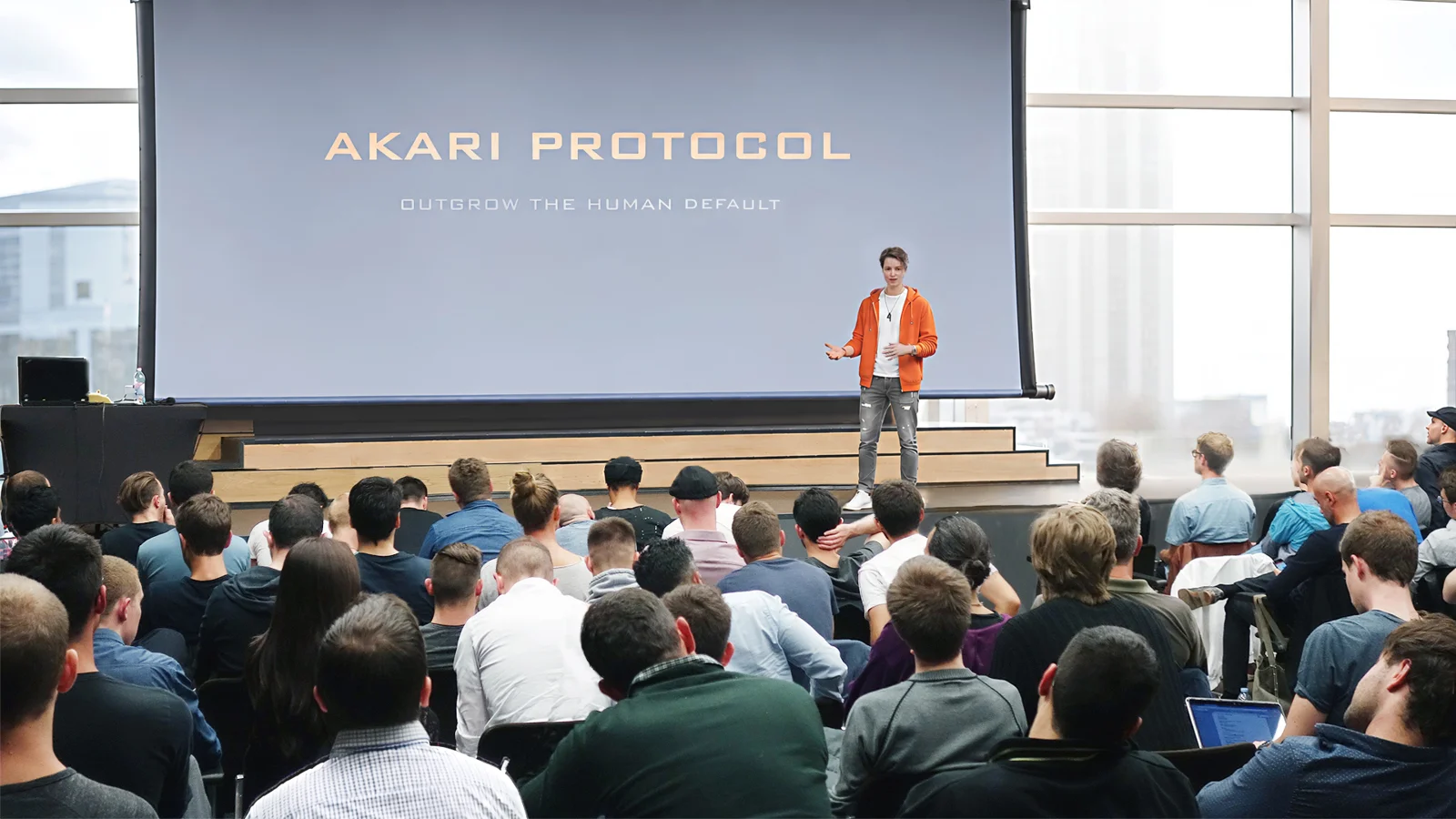 J. H. Tepley speaking on stage in front of an Akari Protocol slide